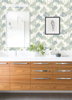 Modern bathroom featuring a wood vanity, white countertop, green plant, and toiletries. A large mirror and framed sketch adorn a wall covered in Lykke Textured Tree Wallpaper by York Wallcoverings. Reflections of a shower appear in the mirror.
