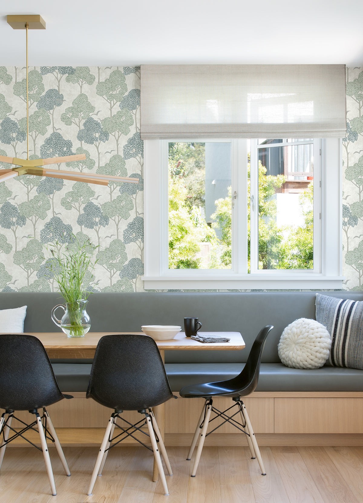 A modern dining nook featuring black chairs, a wooden table, light bench seating, a window overlooking greenery, minimalist decor, and walls adorned with York Wallcoverings’ Lykke Textured Tree Wallpaper.