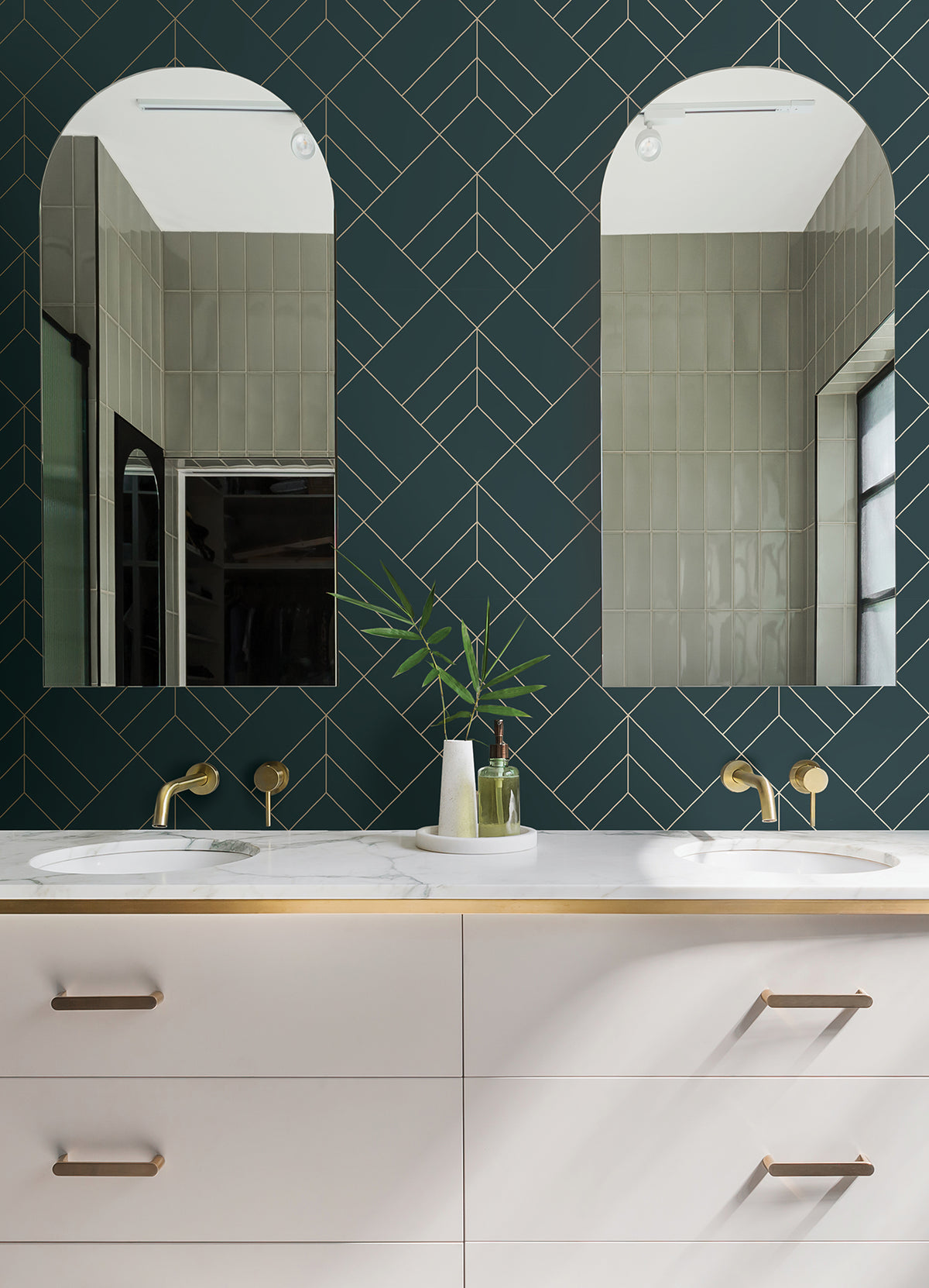 Modern bathroom featuring a double vanity with gold faucets, a white countertop, and two arched mirrors. The backsplash showcases York Wallcoverings’ Sander Geometric Wallpaper in a dark green chevron design, while simple decorative items—including a vase with green leaves—adorn the counter.
