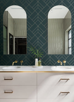 Double bathroom vanity with brass fixtures, white countertop, two modern mirrors, York Wallcoverings Sander Geometric Wallpaper on the wall, and a white vase with green leaves between the sinks. Light reflects from a window on the right.