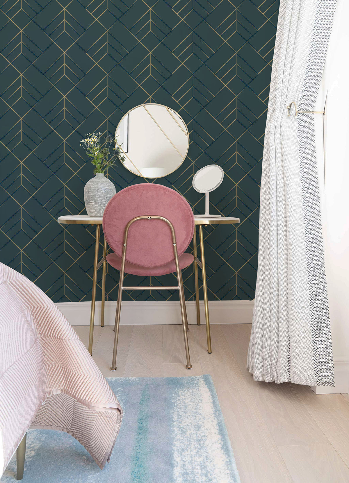 A small vanity with a pink chair, gold legs, round mirrors, and a vase of flowers sits against the Sander Geometric Wallpaper by York Wallcoverings. Cream curtains and a bed with a striped blanket are also visible.