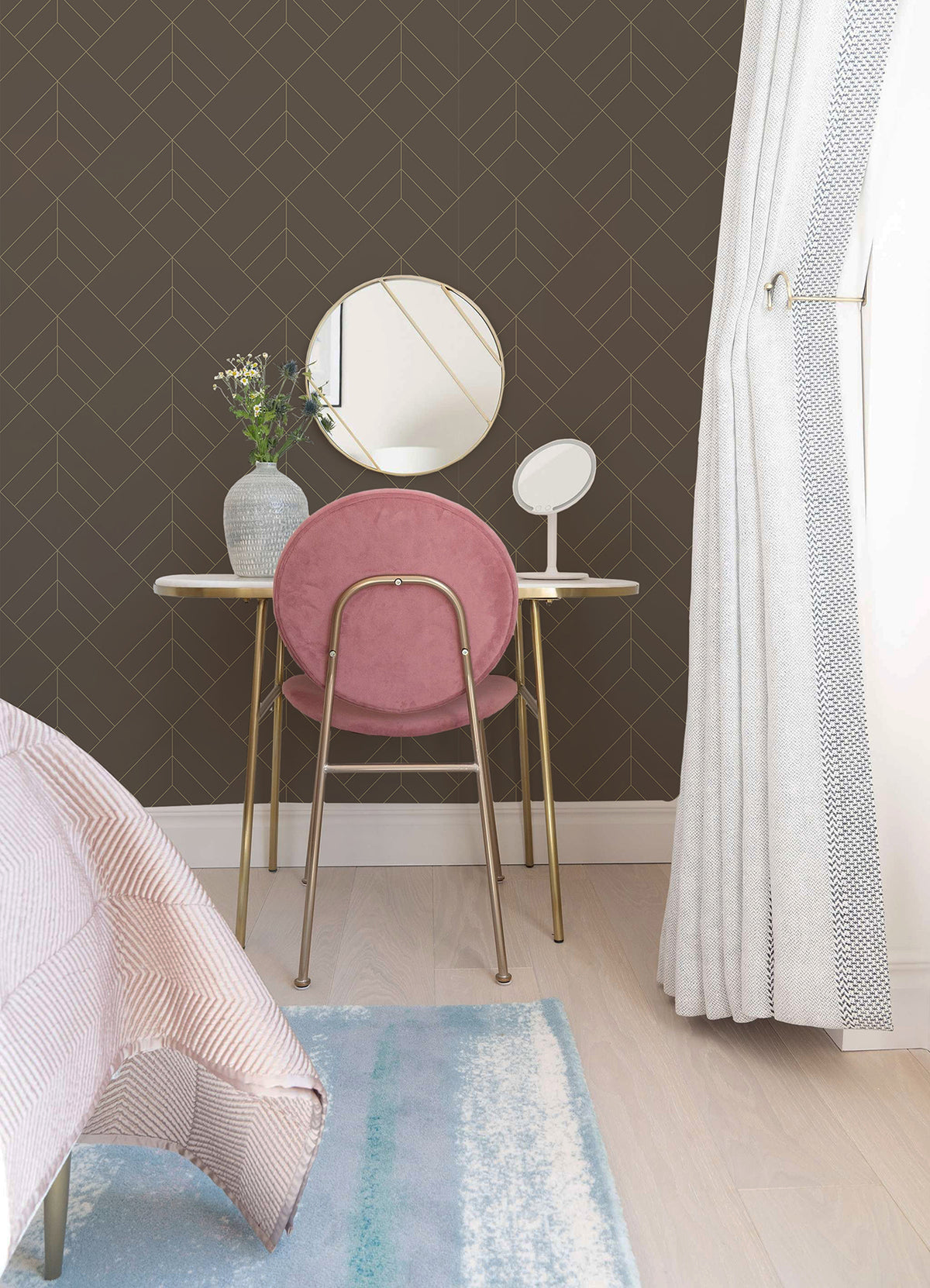 A cozy, modern bedroom features the Sander Geometric Wallpaper by York Wallcoverings as a striking accent behind a vanity set with a pink chair, round mirror, small makeup mirror, and vase of flowers, positioned beside a window dressed in light curtains.