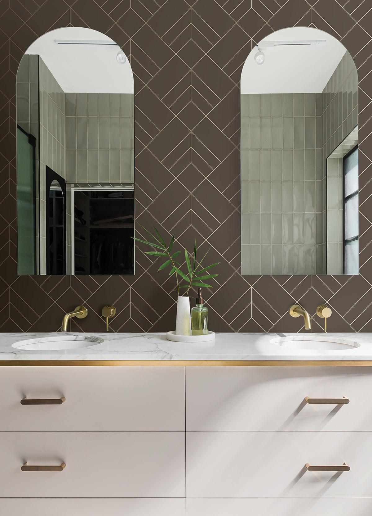 Modern bathroom with two arched mirrors, brass faucets, a white double-sink vanity, and a geometric dark brown Sander Geometric Wallpaper by York Wallcoverings as the backsplash. A small vase with green leaves and toiletries sit on the counter.