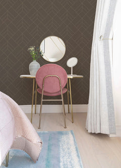 A small vanity table with a round mirror, pink chair, vase of flowers, and table mirror stands against a wall covered in York Wallcoverings’ Sander Geometric Wallpaper. Light curtains and part of a bed and rug are visible in the foreground.