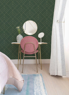 A vanity table with a round mirror, pink chair, small vase with flowers, and accessories sits against a wall adorned with York Wallcoverings’ Sander Geometric Wallpaper next to a window with light curtains. Part of a bed and rug are visible.
