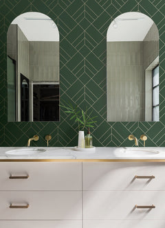 Modern bathroom featuring two arched mirrors, a green Sander Geometric Wallpaper by York Wallcoverings on the wall, double sinks with brass faucets, and a white vanity adorned with gold accents. A vase with green leaves is placed between the sinks.