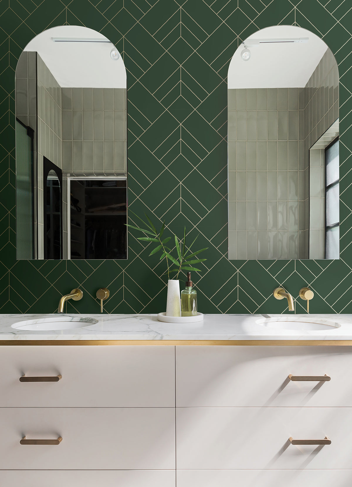 Modern bathroom featuring Sander Geometric Wallpaper by York Wallcoverings on the walls, two arched mirrors, brass fixtures, and a white double-sink vanity with gold-trimmed drawers. A small vase with greenery sits on the countertop.