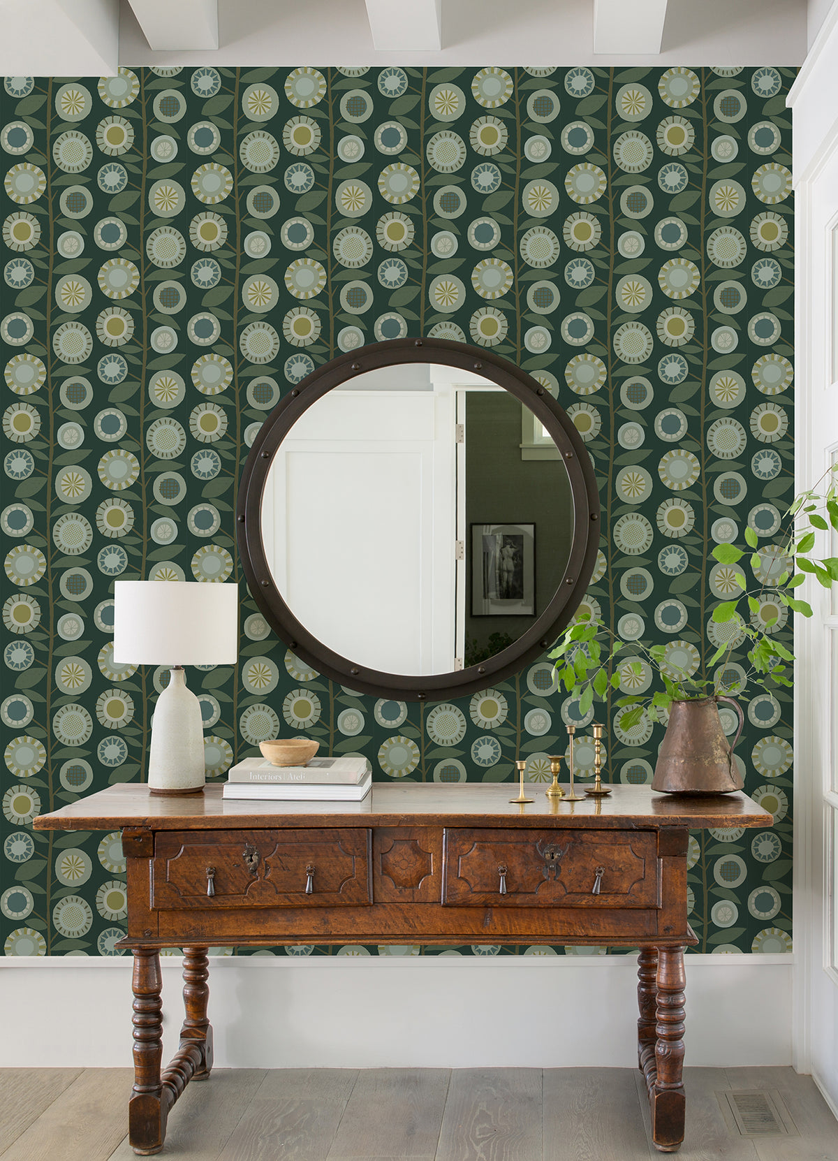 A wooden console table sits against a wall adorned with York Wallcoverings’ Sisu Floral Geometric Wallpaper in green, topped with a lamp, books, bowl, and vase. A round mirror is mounted above, reflecting a white door and framed art.