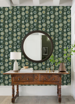 A wooden console table sits against a wall adorned with York Wallcoverings’ Sisu Floral Geometric Wallpaper in green, topped with a lamp, books, bowl, and vase. A round mirror is mounted above, reflecting a white door and framed art.