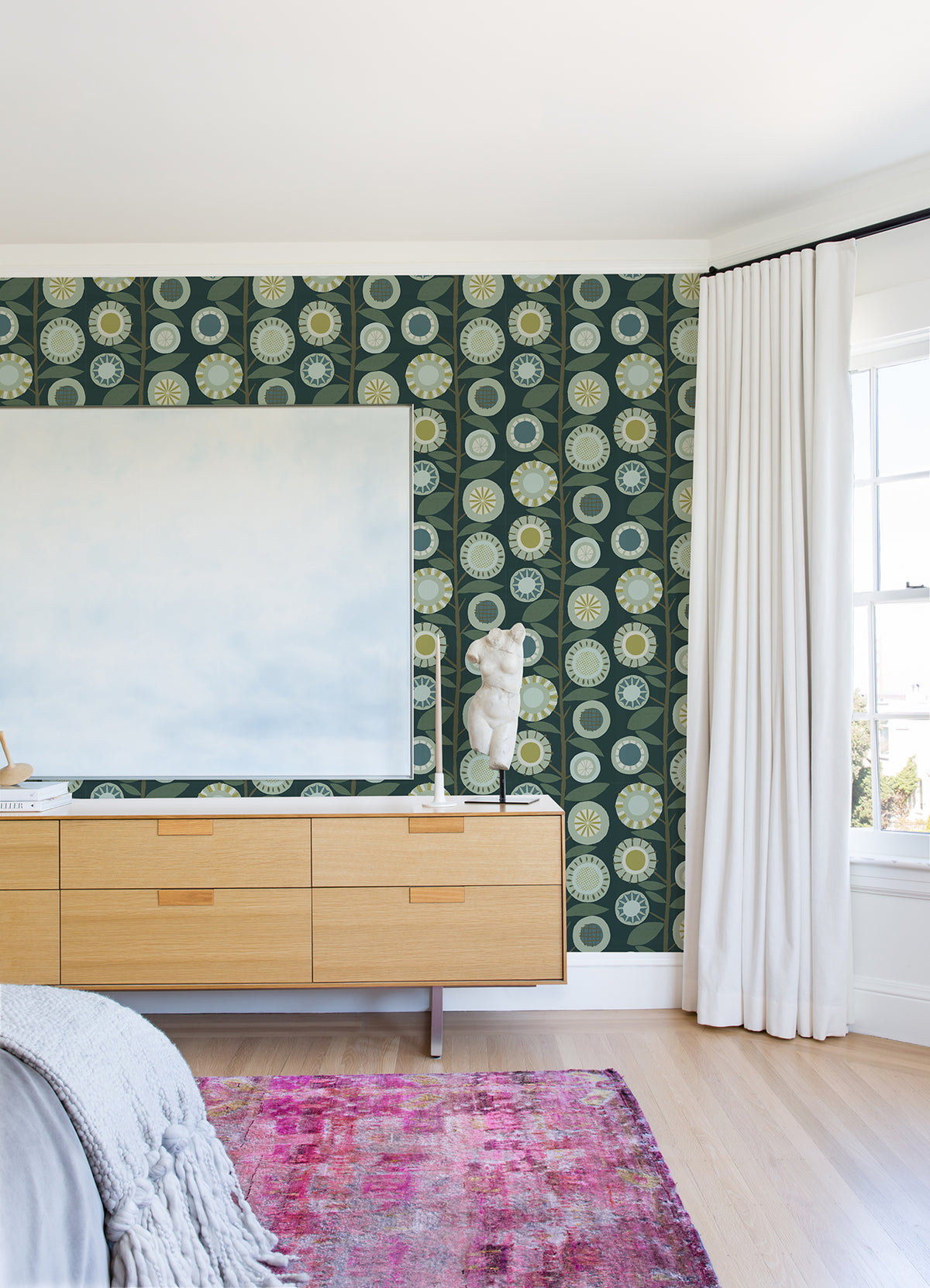 A modern bedroom featuring York Wallcoverings’ Sisu Floral Geometric Wallpaper, a light wood dresser, an abstract statue, a large blank canvas, white curtains, and a pink rug on a light wooden floor with sunlight streaming through the window.