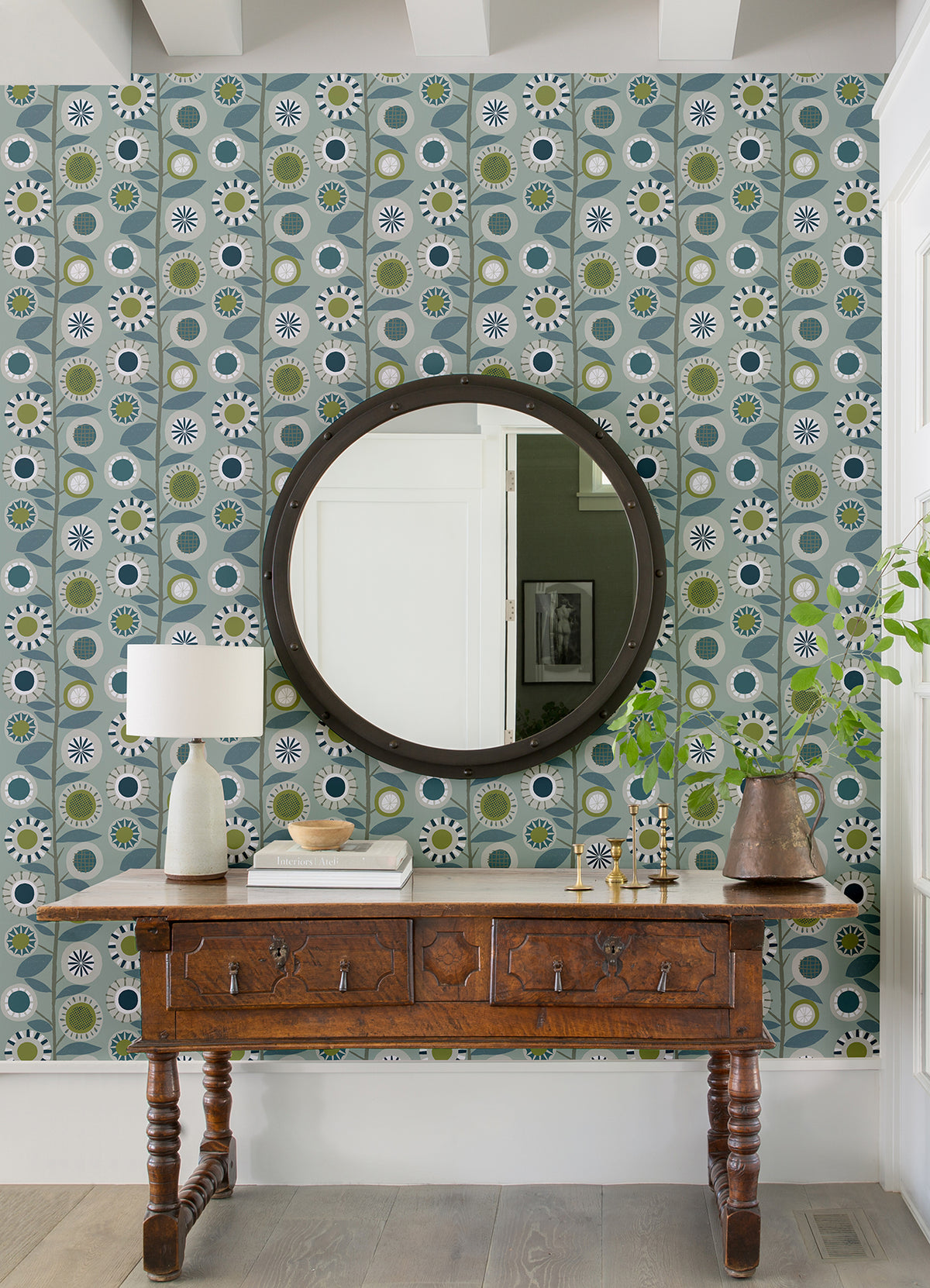 A wooden table adorned with a lamp, books, and decorative accents is set against York Wallcoverings’ Sisu Floral Geometric Wallpaper in blue. Above the table hangs a round mirror reflecting a bright white wall and photograph, while a vase with a leafy plant decorates the right end of the tabletop.
