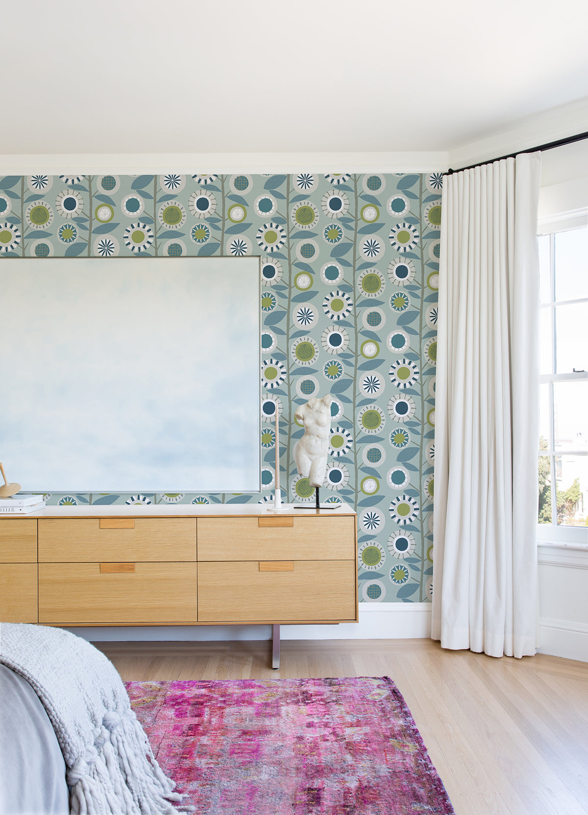 A bedroom features a light wood dresser, abstract art, white curtains, a pink rug, and a small sculpture on the dresser. The walls are adorned with Sisu Floral Geometric Wallpaper by York Wallcoverings, while sunlight pours in through a large window.
