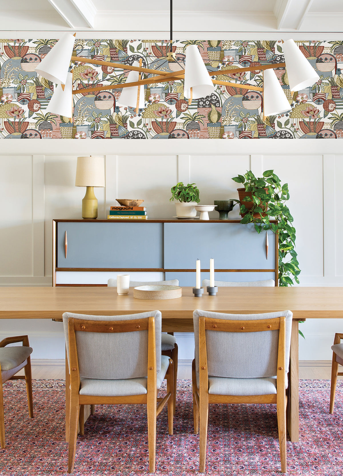 A dining room with four wooden chairs, a table, a mid-century cabinet, plants, candles, and a gold lamp features Fika Blissful Birds & Blooms Wallpaper by York Wallcoverings above white paneling. A modern white chandelier and a red patterned rug complete the space.