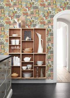 Open wooden shelves displaying dishes and glassware stand against the Fika Blissful Birds & Blooms Wallpaper by York Wallcoverings, which features colorful, vintage-inspired floral and fruit motifs. A gray kitchen island and an arched doorway are also visible.