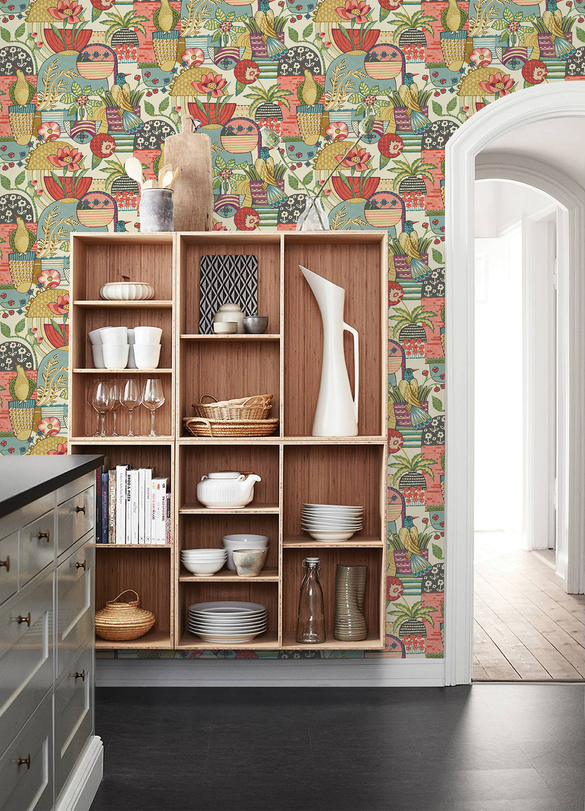 A wooden shelving unit with dishes and glassware sits in a kitchen, where the wall behind features York Wallcoverings’ Fika Blissful Birds & Blooms Wallpaper. An arched doorway opens to a bright hallway with wood floors.