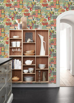 A wooden shelving unit with dishes and glassware sits in a kitchen, where the wall behind features York Wallcoverings’ Fika Blissful Birds & Blooms Wallpaper. An arched doorway opens to a bright hallway with wood floors.