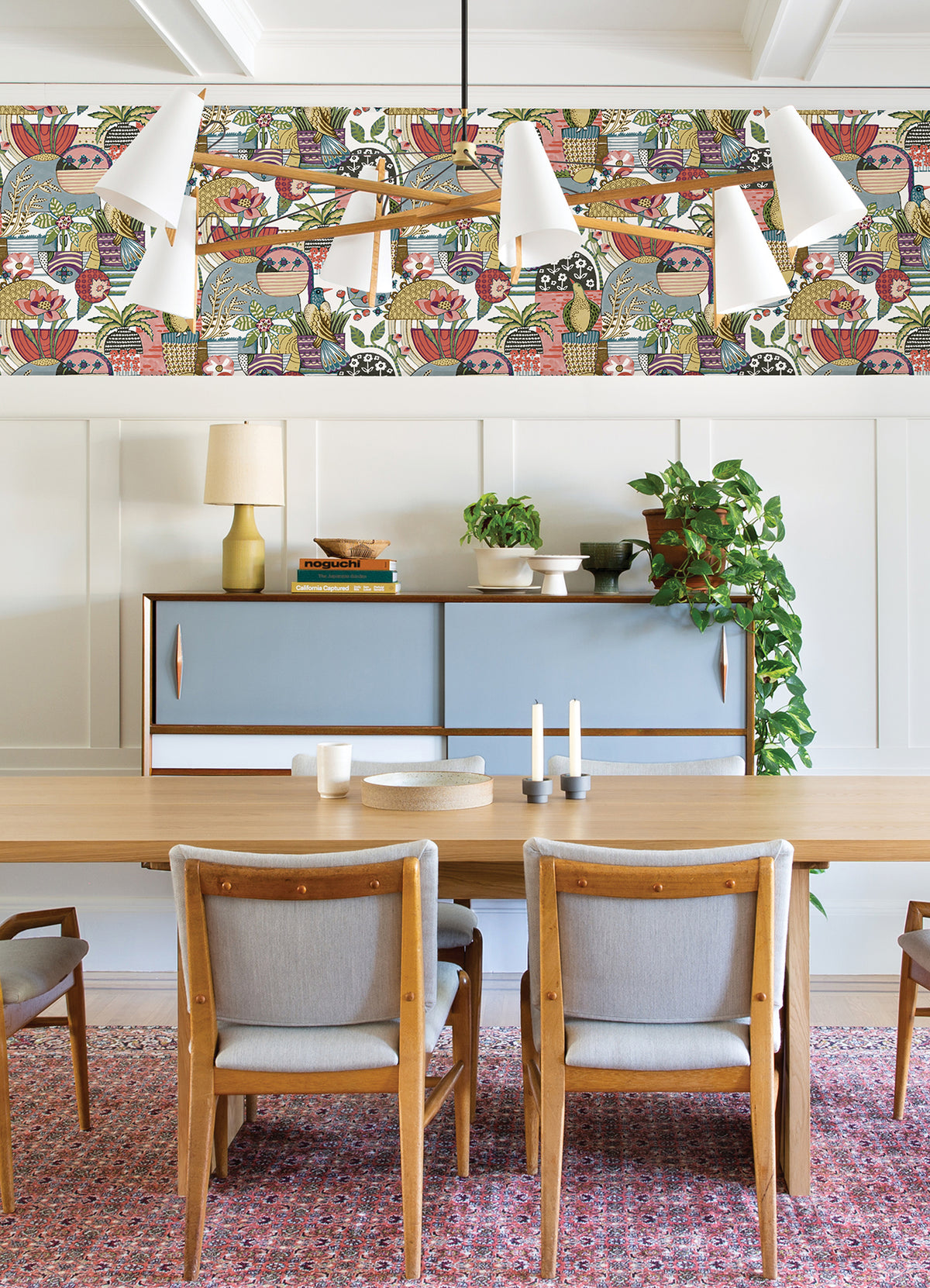 Dining room with a wooden table, gray chairs, sideboard, leafy plant, candlesticks, and Fika Blissful Birds & Blooms Wallpaper by York Wallcoverings. A modern chandelier hangs above the table over a red-patterned rug.