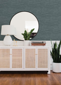 A modern sideboard with a white lamp, potted plants, and a record player sits beneath a round mirror on a wall adorned with Malin Faux Grasscloth Wallpaper by York Wallcoverings. The floor is wooden.