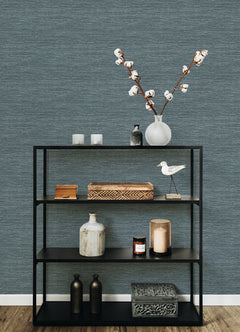 A black metal shelf displays decorative items—a vase filled with cotton branches, candles, boxes, jars, a ceramic bird, and a patterned box—set against a York Wallcoverings Malin Faux Grasscloth Wallpaper in blue-gray tones atop a wooden floor.