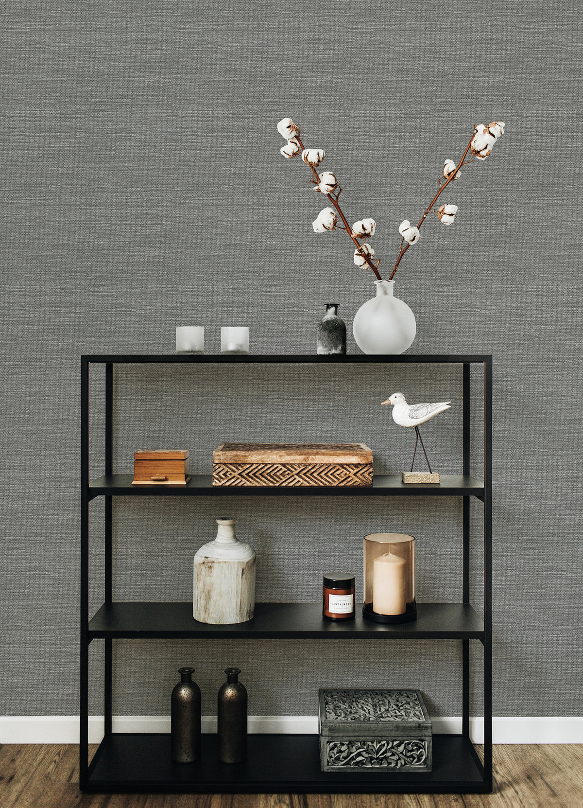 A black metal shelf displays decorative items—a vase with cotton stems, candles, boxes, bottles, a bird figurine, and a patterned box—set against a wall adorned with York Wallcoverings’ Malin Faux Grasscloth Wallpaper and a wooden floor.
