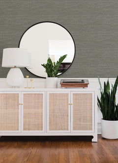 A white sideboard with rattan doors holds a lamp, gold candlesticks, plant, and record player. Above it hangs a round mirror on walls adorned with Malin Faux Grasscloth Wallpaper by York Wallcoverings, while a tall plant in a white pot sits on the wood floor beside the cabinet.
