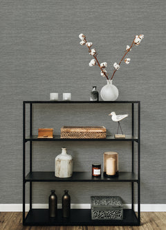 A black metal shelf stands against a wall adorned with Malin Faux Grasscloth Wallpaper by York Wallcoverings, displaying neatly arranged decorative items: a vase with cotton branches, candles, small boxes, bottles, a bird figurine, and a patterned box on its three shelves.