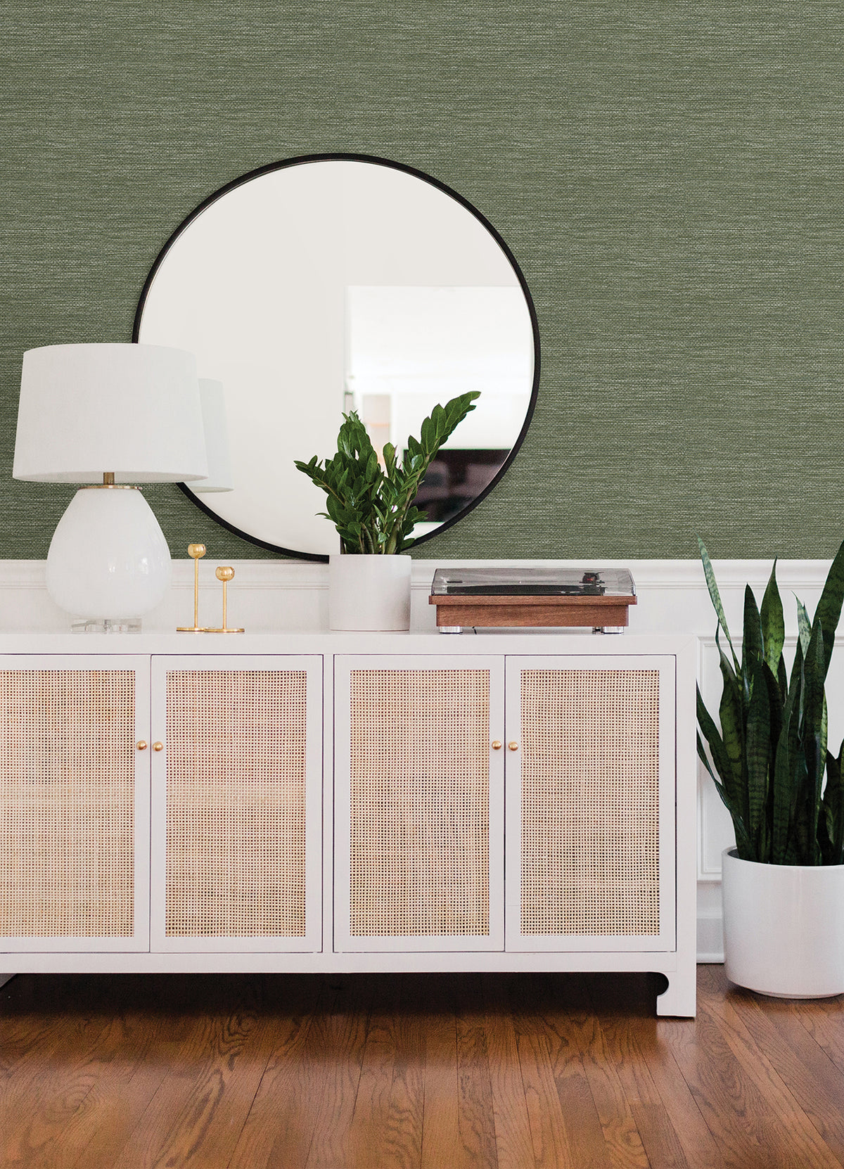 A white sideboard with cane doors sits on a wooden floor, topped with a white lamp, green plants, and decor. A round mirror hangs above against a wall covered in Malin Faux Grasscloth Wallpaper by York Wallcoverings.