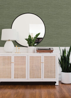 A white cabinet with cane doors holds a white lamp, small green plant, and record player. A large round mirror hangs above, reflecting the room’s Malin Faux Grasscloth Wallpaper by York Wallcoverings. A tall potted plant sits on the wooden floor nearby.