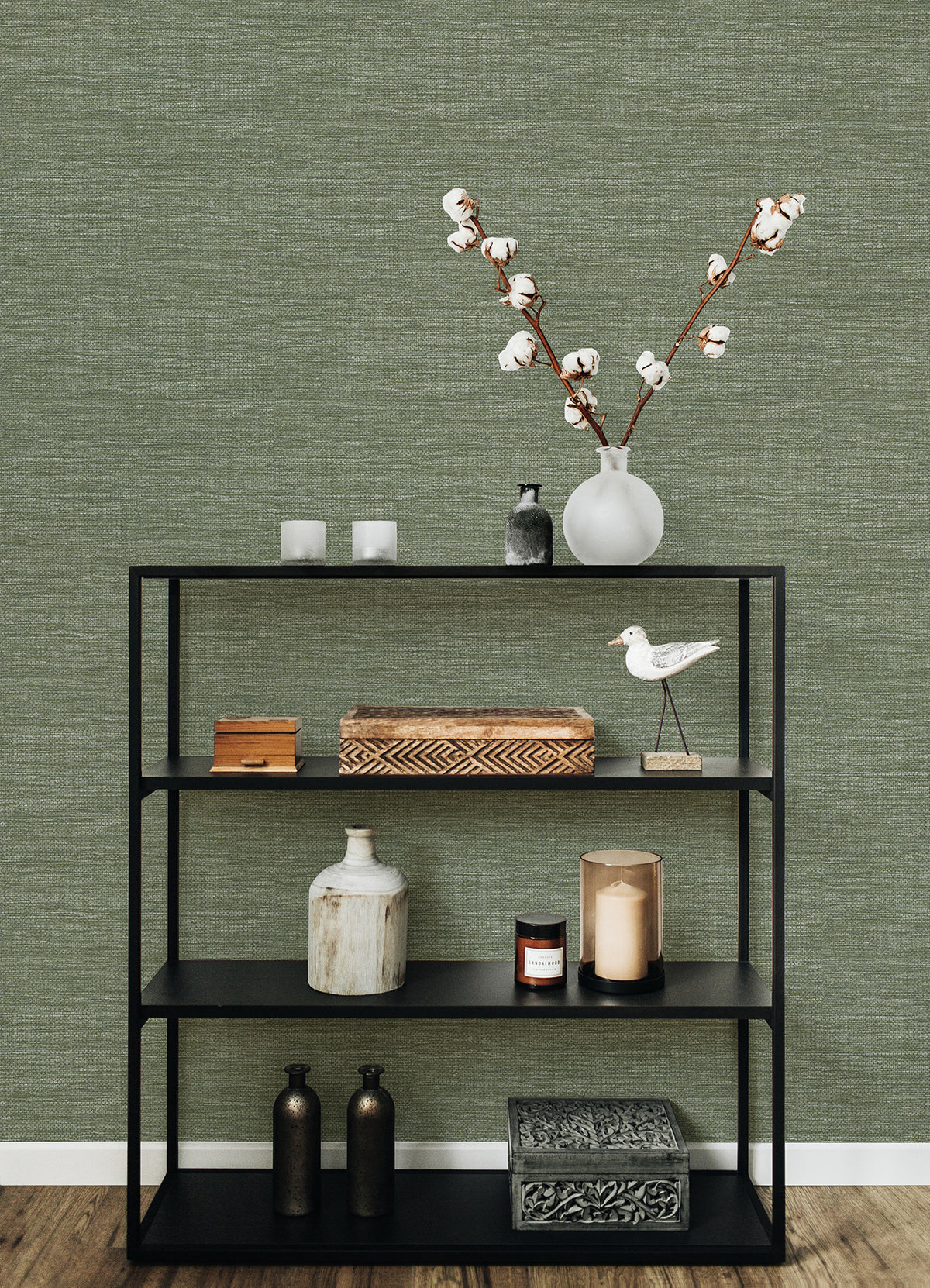 A black shelving unit holds decorative objects—a vase with cotton stems, candles, boxes, bottles, and a white bird figurine—set against a wooden floor and a textured green wall covered in Malin Faux Grasscloth Wallpaper by York Wallcoverings.