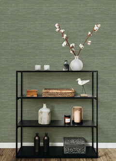 A black shelving unit holds decorative objects—a vase with cotton stems, candles, boxes, bottles, and a white bird figurine—set against a wooden floor and a textured green wall covered in Malin Faux Grasscloth Wallpaper by York Wallcoverings.
