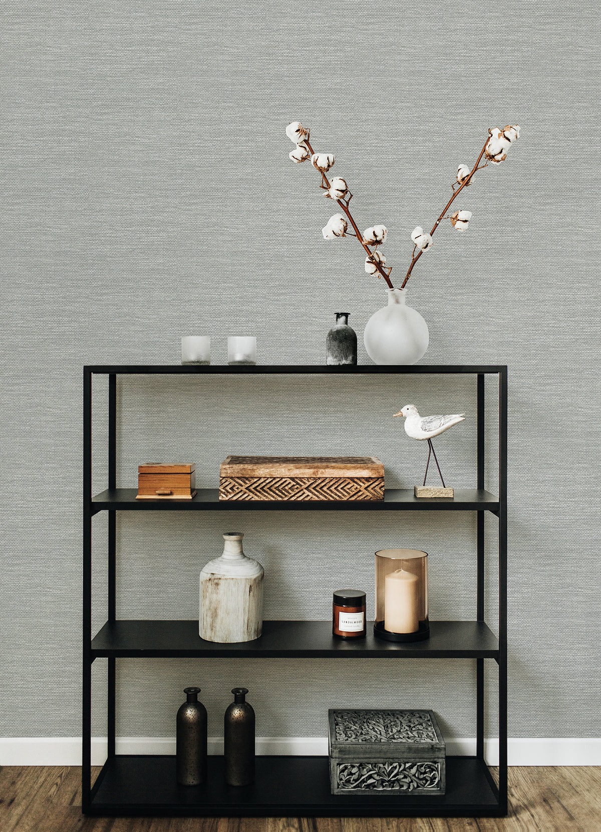 A black metal shelf displaying decorative items—vases, a wooden box, candles, a ceramic bird, and a branch with cotton in a white vase—stands out against walls adorned with York Wallcoverings’ Malin Faux Grasscloth Wallpaper.