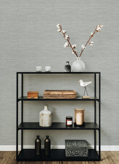 A black metal shelf displaying decorative items—vases, a wooden box, candles, a ceramic bird, and a branch with cotton in a white vase—stands out against walls adorned with York Wallcoverings’ Malin Faux Grasscloth Wallpaper.