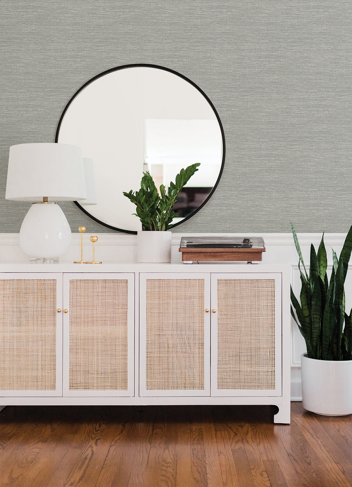 Malin Faux Grasscloth Wallpaper by York Wallcoverings creates a stylish backdrop for a white sideboard with rattan doors, which displays a lamp, two gold candleholders, a plant, and a record player. A round mirror hangs above the sideboard, while a tall potted plant stands on the wood floor to the right.