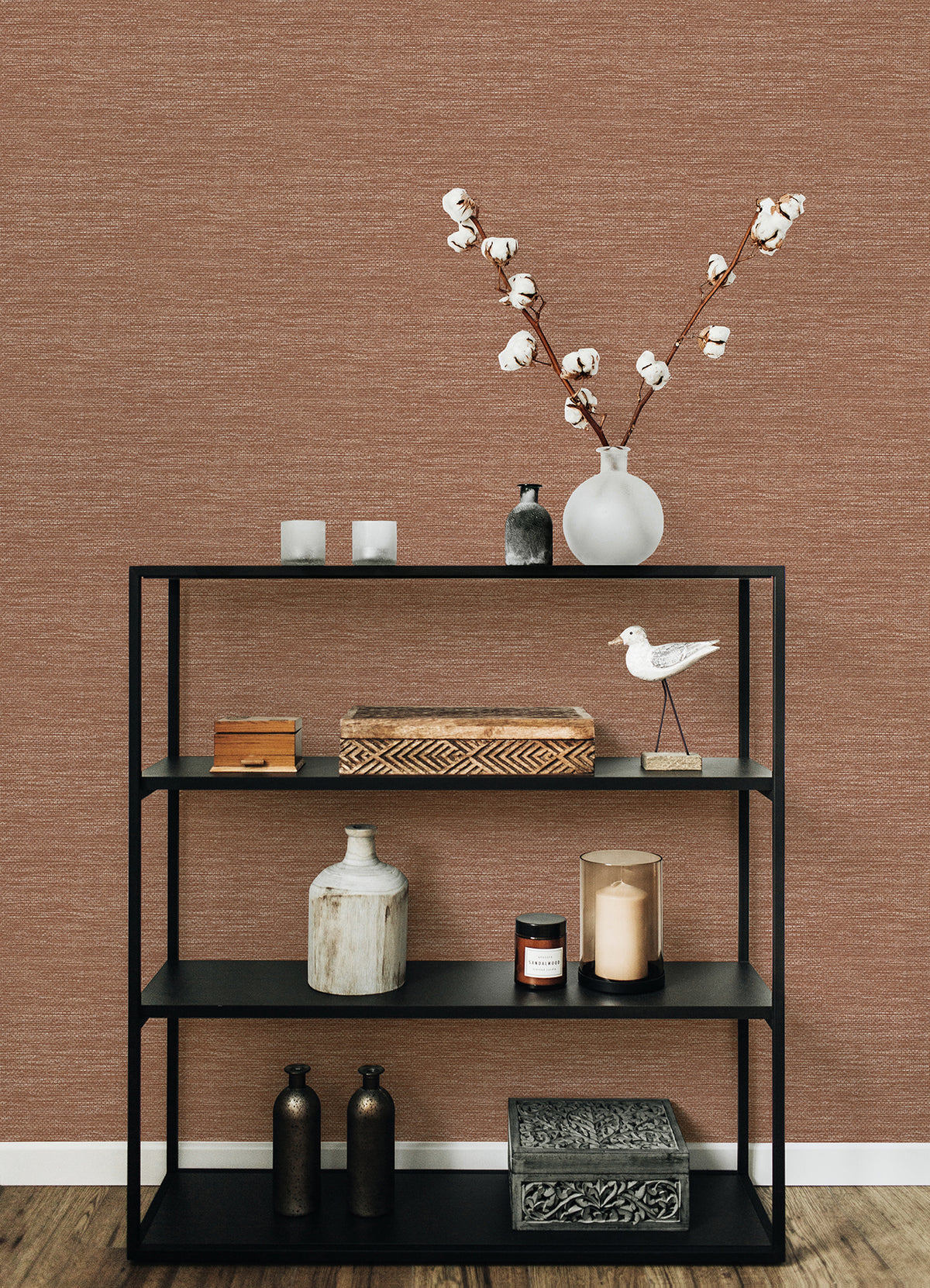 A black metal shelf styled with decorative items—including vases, candles, boxes, and a white ceramic bird—sits in front of a wall featuring Malin Faux Grasscloth Wallpaper by York Wallcoverings. On the top shelf, a glass vase filled with cotton stems stands out against the wallpaper’s rich brown texture.