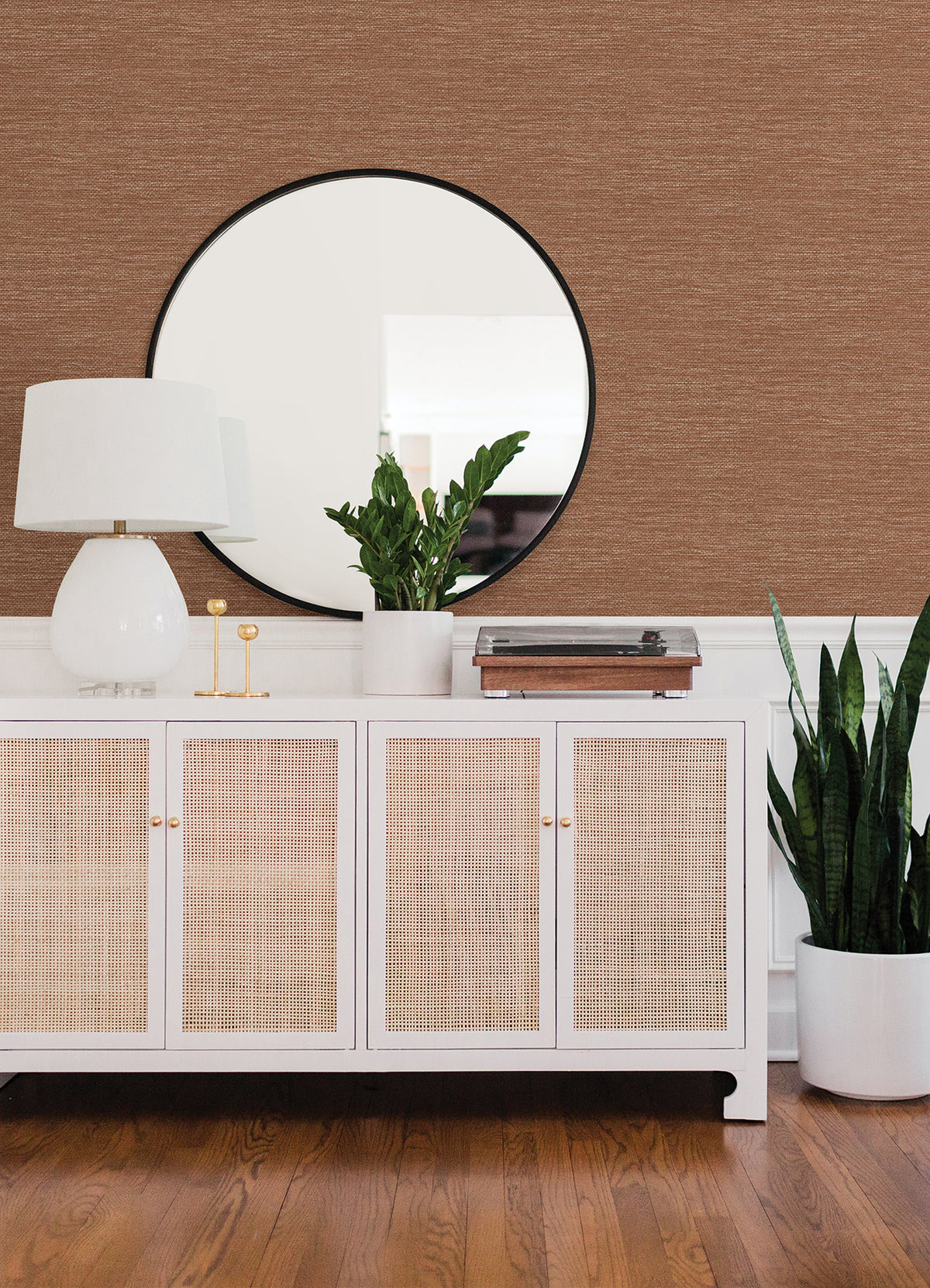 A white sideboard with rattan doors holds a white lamp, a round mirror, a potted plant, and decor items. A tall plant in a white pot stands on a wooden floor against a wall covered in Malin Faux Grasscloth Wallpaper by York Wallcoverings.