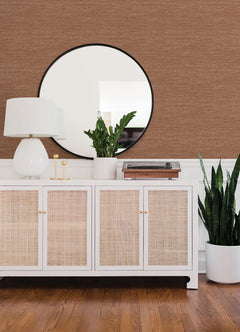 A white sideboard with rattan doors holds a white lamp, a round mirror, a potted plant, and decor items. A tall plant in a white pot stands on a wooden floor against a wall covered in Malin Faux Grasscloth Wallpaper by York Wallcoverings.