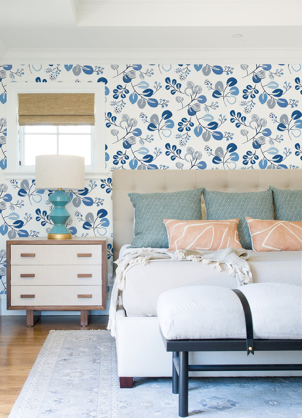 Bedroom with York Wallcoverings’ Jonah Leaf Trail Wallpaper in blue and white, a tufted bed adorned with patterned pillows, a white bench, a nightstand featuring a turquoise lamp, and a small window with a woven shade.
