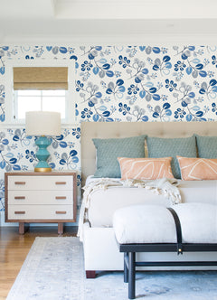 Bedroom with York Wallcoverings’ Jonah Leaf Trail Wallpaper in blue and white, a tufted bed adorned with patterned pillows, a white bench, a nightstand featuring a turquoise lamp, and a small window with a woven shade.