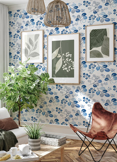 A cozy living room featuring York Wallcoverings’ Jonah Leaf Trail Wallpaper, three framed botanical prints, wicker ceiling lights, a green plant, a brown butterfly chair, and a small table adorned with potted plants. Sunlight streams through sheer curtains.