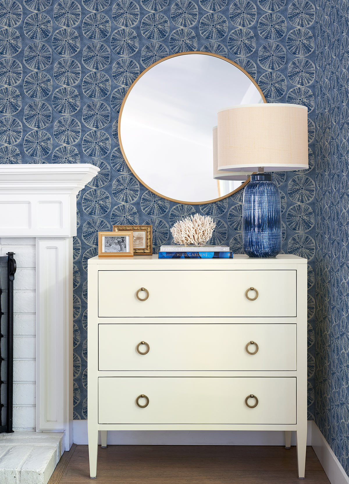 A white dresser with four circular handles holds a blue glass lamp, white coral decor, books, and framed photos. Above it is a round mirror. The background features Chesapeake Sea Biscuit Sand Dollar Wallpaper by York Wallcoverings in a blue pattern and a white fireplace.