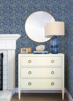 A white dresser with four circular handles holds a blue glass lamp, white coral decor, books, and framed photos. Above it is a round mirror. The background features Chesapeake Sea Biscuit Sand Dollar Wallpaper by York Wallcoverings in a blue pattern and a white fireplace.