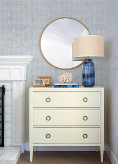 A cream dresser with three drawers sits beside a white fireplace, set against Chesapeake Wellen Abstract Rope Wallpaper by York Wallcoverings. On top are a blue lamp, a white coral, books, and framed photos. A round mirror hangs above, reflecting part of the room.