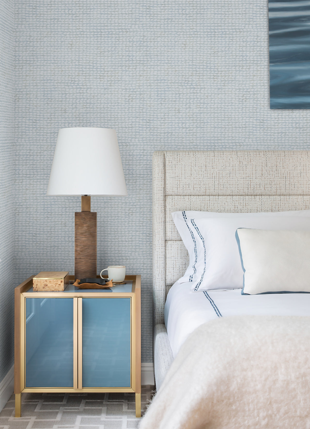 A modern bedroom featuring a light upholstered bed with white bedding and a blue-accented pillow is complemented by a gold nightstand topped with a brown lamp, a book, and a cup. The wall is finished in Chesapeake Wellen Abstract Rope Wallpaper from York Wallcoverings in a light blue hue, adding texture to the space. Part of a painting is also visible.