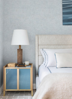 A modern bedroom featuring a light upholstered bed with white bedding and a blue-accented pillow is complemented by a gold nightstand topped with a brown lamp, a book, and a cup. The wall is finished in Chesapeake Wellen Abstract Rope Wallpaper from York Wallcoverings in a light blue hue, adding texture to the space. Part of a painting is also visible.