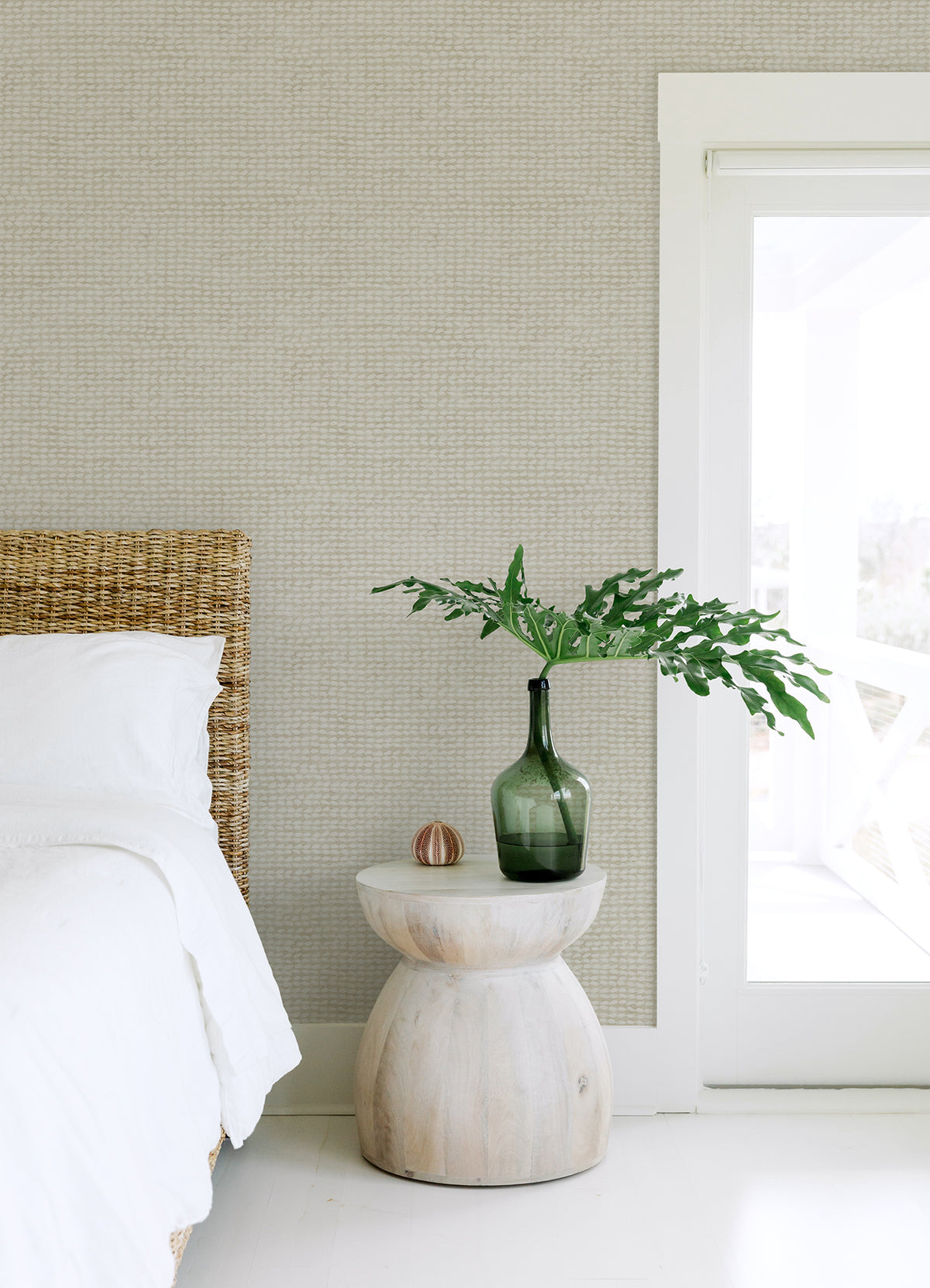 A minimalist bedroom featuring Chesapeake Wellen Abstract Rope Wallpaper by York Wallcoverings, a wicker bed with white bedding, a round wooden side table, a green glass vase with leafy branches, a small decor object, and a glass door letting in natural light.