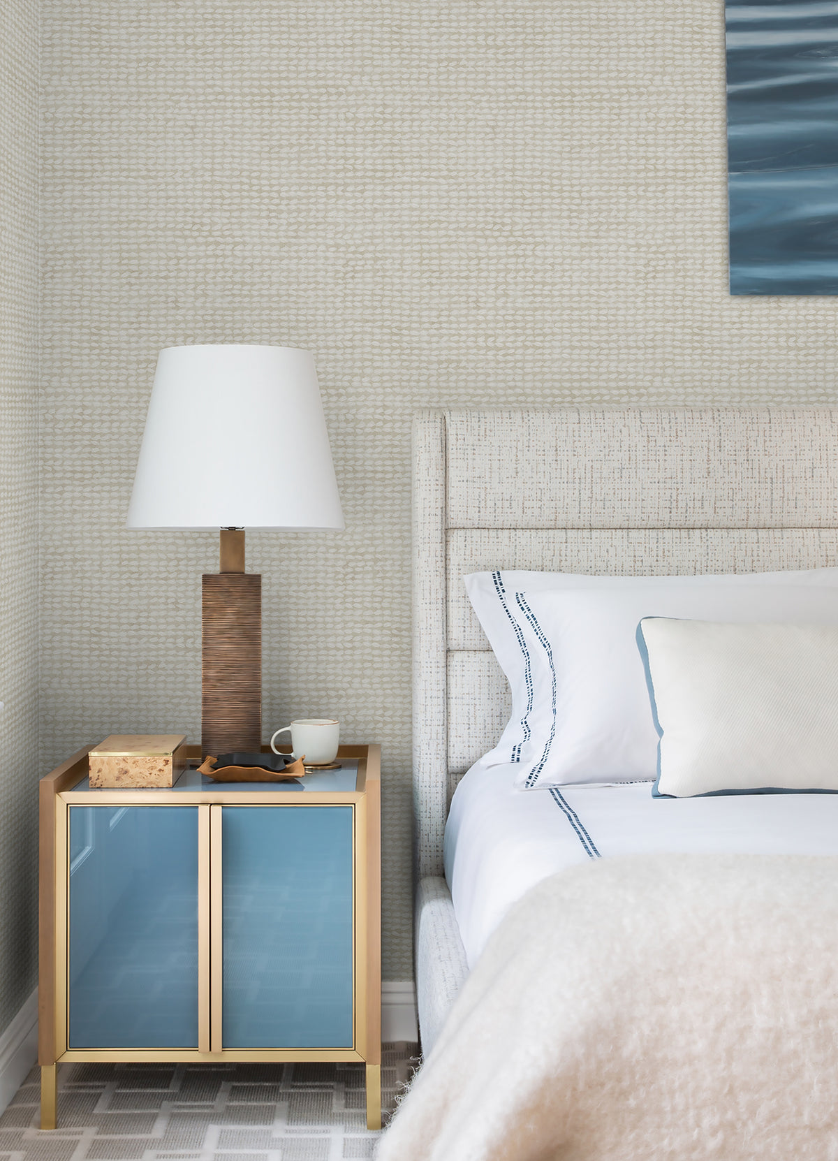 A modern bedroom featuring a beige upholstered bed with white and blue linens, a wooden nightstand with blue doors, a lamp, a mug, and a book, all set against Chesapeake Wellen Abstract Rope Wallpaper by York Wallcoverings.