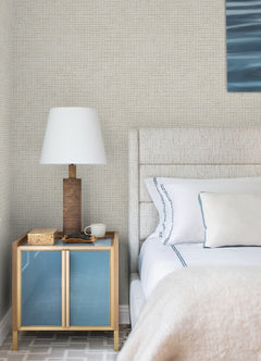 A modern bedroom featuring a beige upholstered bed with white and blue linens, a wooden nightstand with blue doors, a lamp, a mug, and a book, all set against Chesapeake Wellen Abstract Rope Wallpaper by York Wallcoverings.