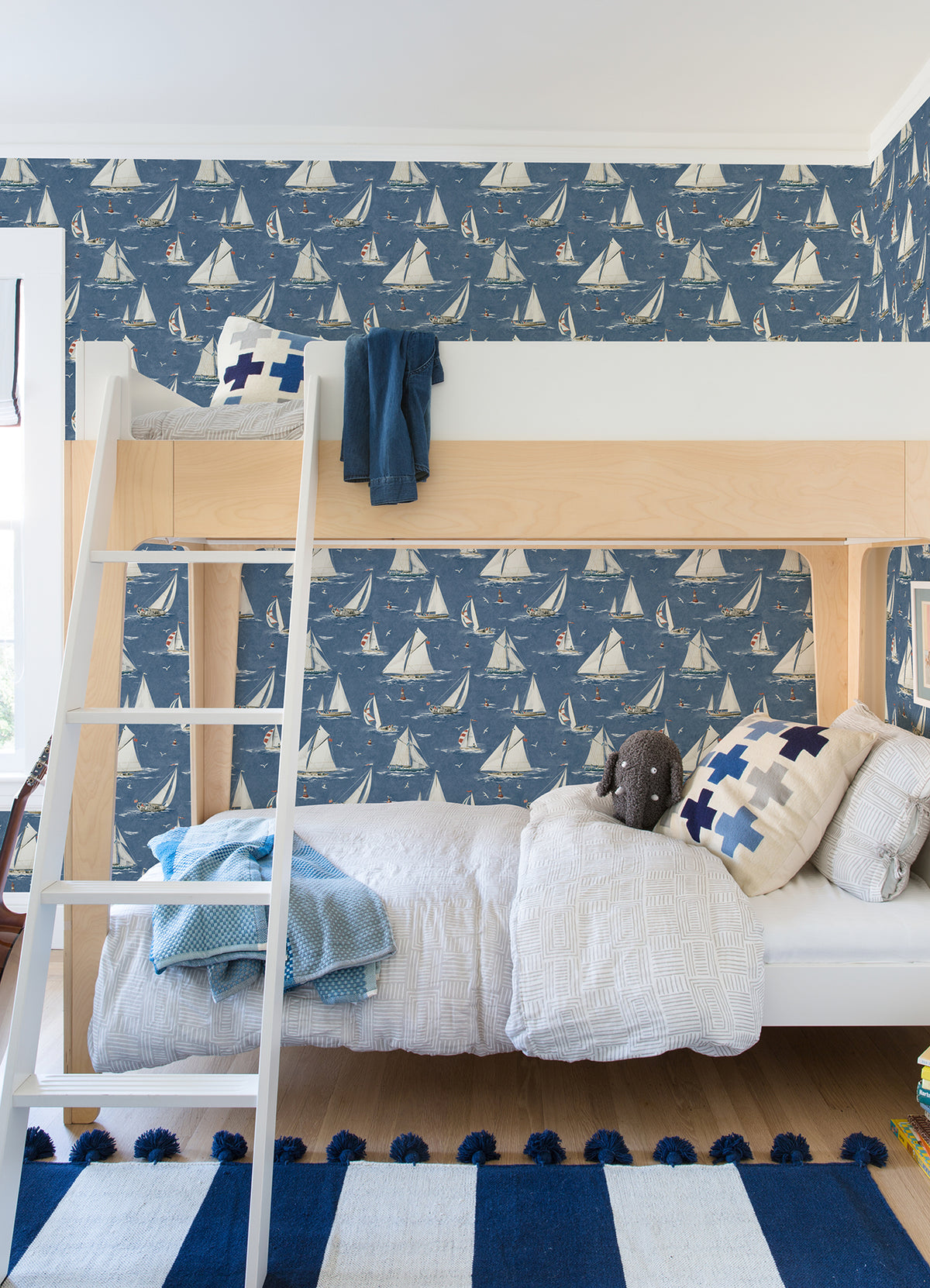 A cozy kids bedroom featuring a wooden bunk bed, Chesapeake Leeward Sailboat Wallpaper by York Wallcoverings, blue and white bedding, plush toys, and a striped blue rug on a wooden floor.
