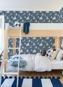 A cozy kids bedroom featuring a wooden bunk bed, Chesapeake Leeward Sailboat Wallpaper by York Wallcoverings, blue and white bedding, plush toys, and a striped blue rug on a wooden floor.