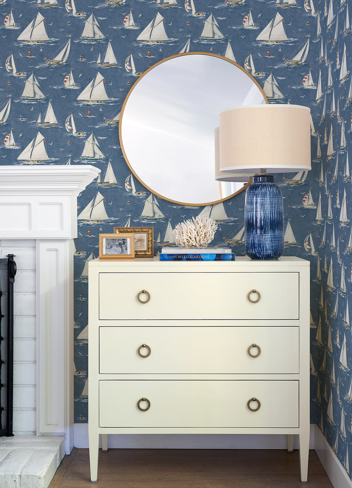 A white dresser with three drawers sits against Chesapeake Leeward Sailboat Wallpaper by York Wallcoverings. A round mirror, a blue glass lamp, coral decor, and framed photos rest on top. Part of a white fireplace is visible on the left.